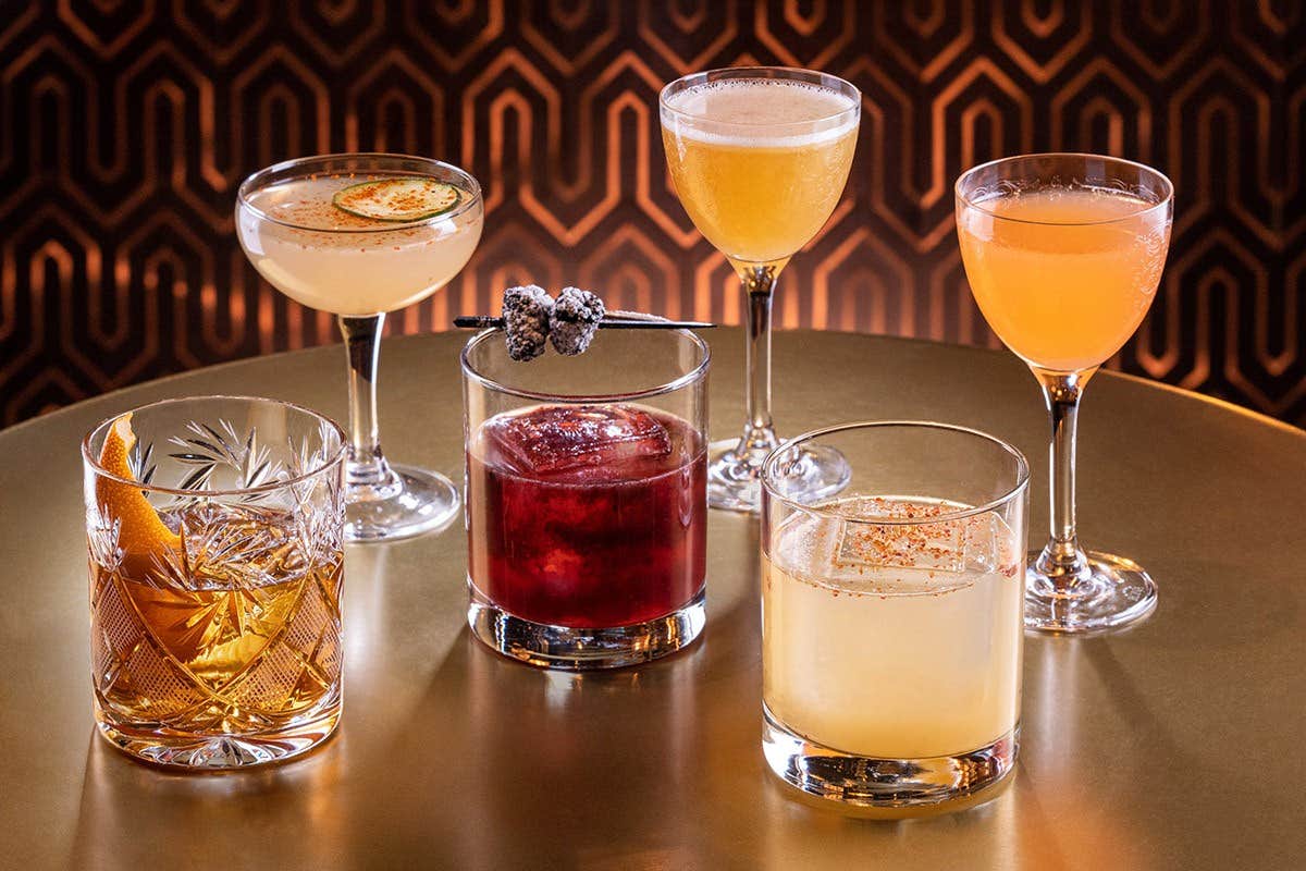 A variety of six colorful cocktails on a round, gold table with a geometric-patterned background.