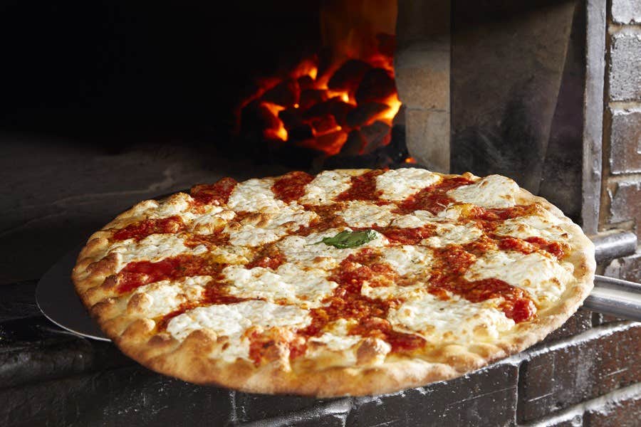 A pizza with a crispy crust, melted cheese, and tomato sauce is being taken out of a brick oven with a paddle.