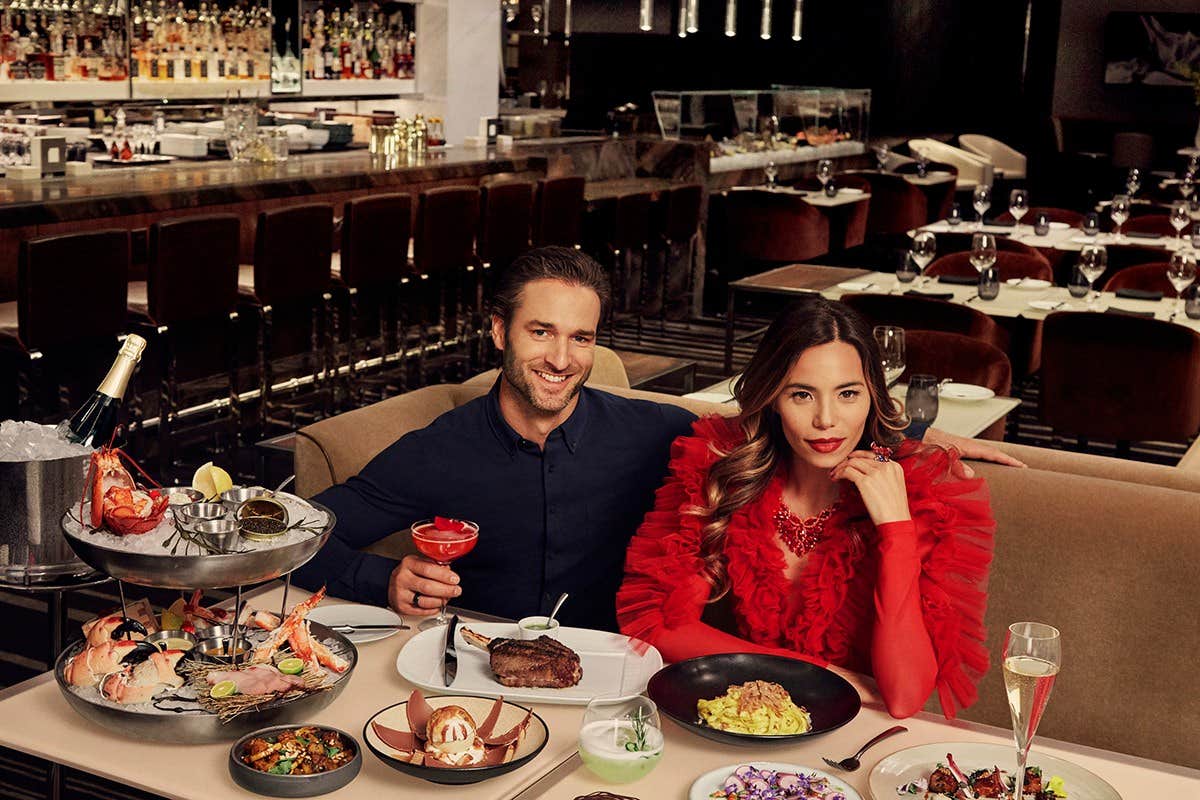 A man and woman enjoy fine dining at an upscale restaurant, with an array of dishes and drinks on the table.