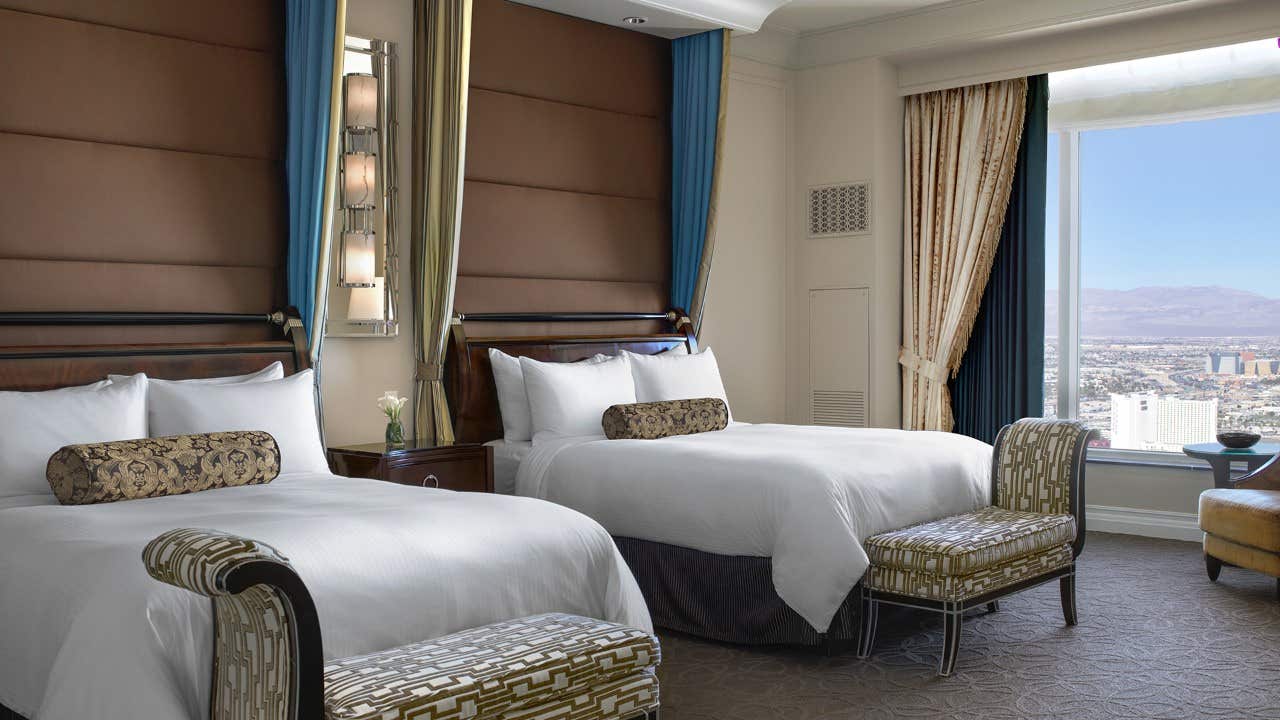 Luxury hotel room with two double beds, elegant decor, and a view of the city and mountains through a large window.