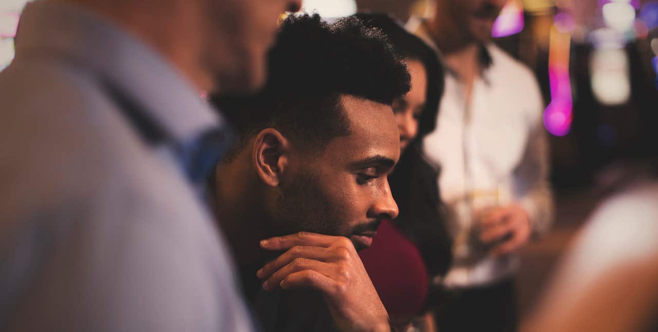 A man with short hair looks thoughtfully downwards; blurred people and lights are in the background.