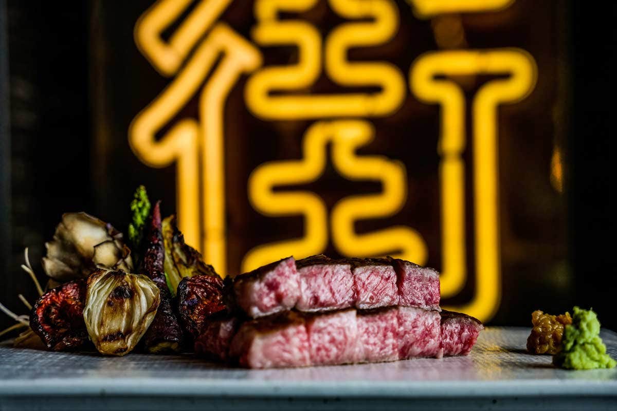 Slices of seared steak served with vegetables, set against a backdrop of yellow neon lights.