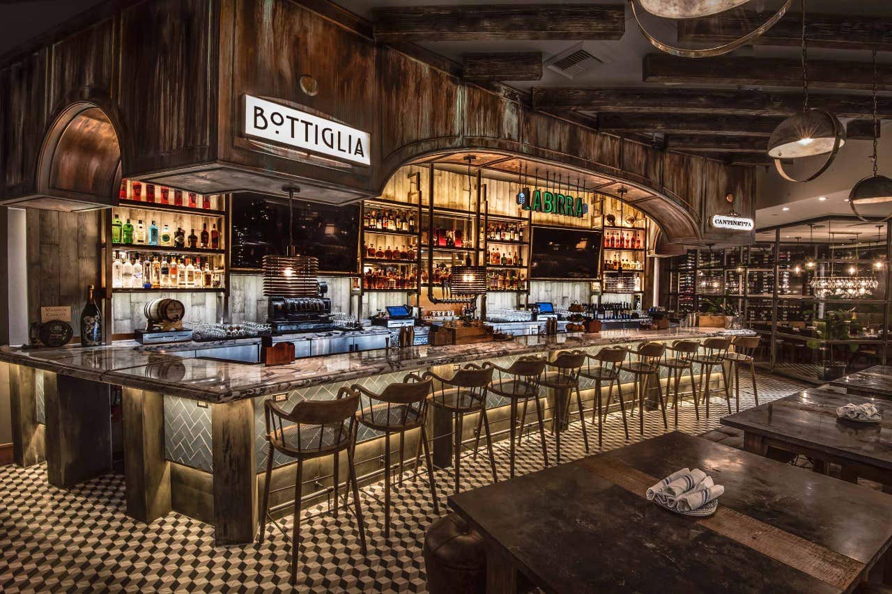 Rustic bar interior with wooden beams, illuminated shelves, a long marble counter, and high stools. Sign reads "Bottiglia.