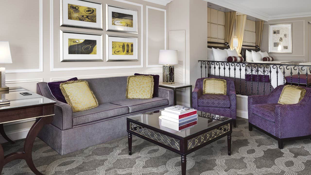 Elegant hotel suite with a sofa, two armchairs, coffee table, desk, and bed in a separate elevated area.