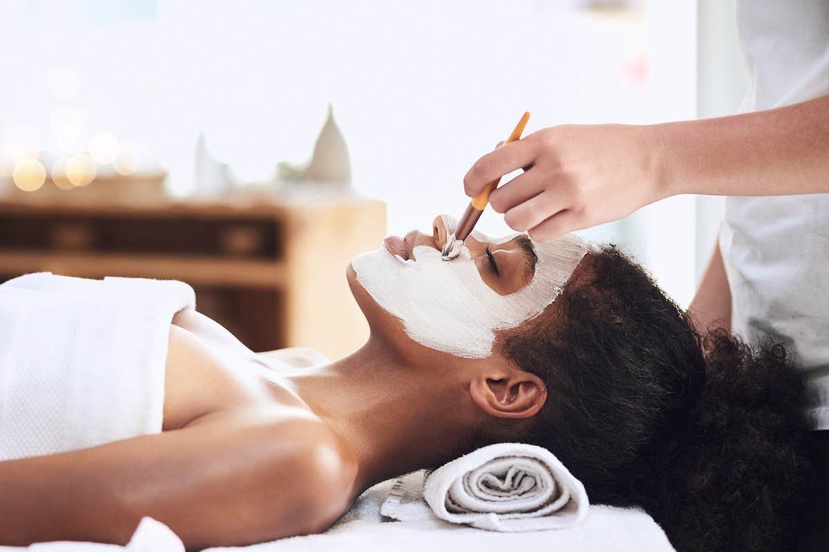 Person receiving a facial treatment, with white cream being applied to their face while lying on a massage table.