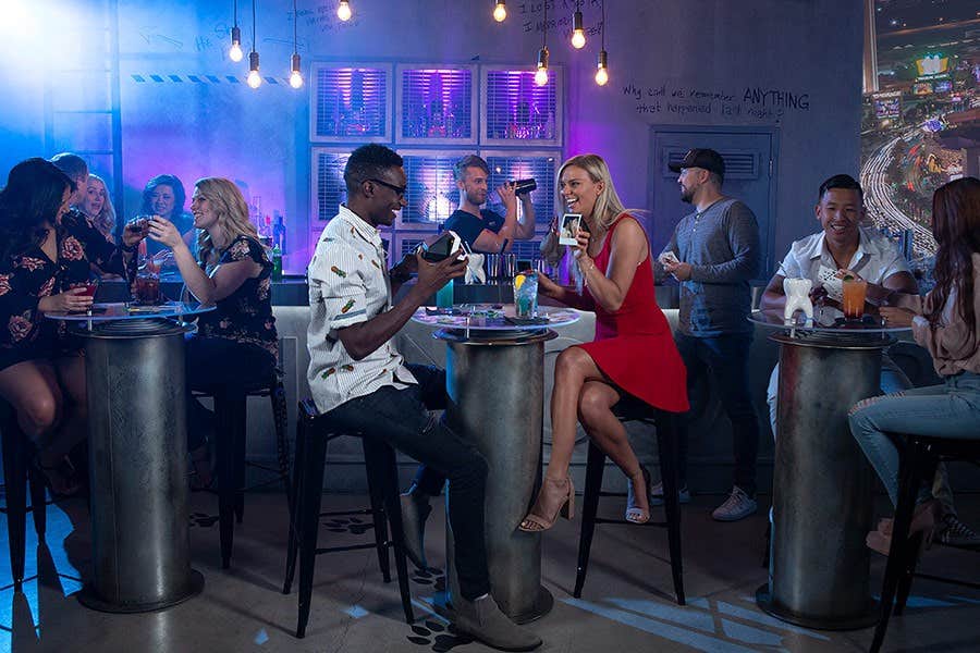 A lively bar scene with people chatting and taking photos, some seated at high tables, and a neon-lit backdrop.