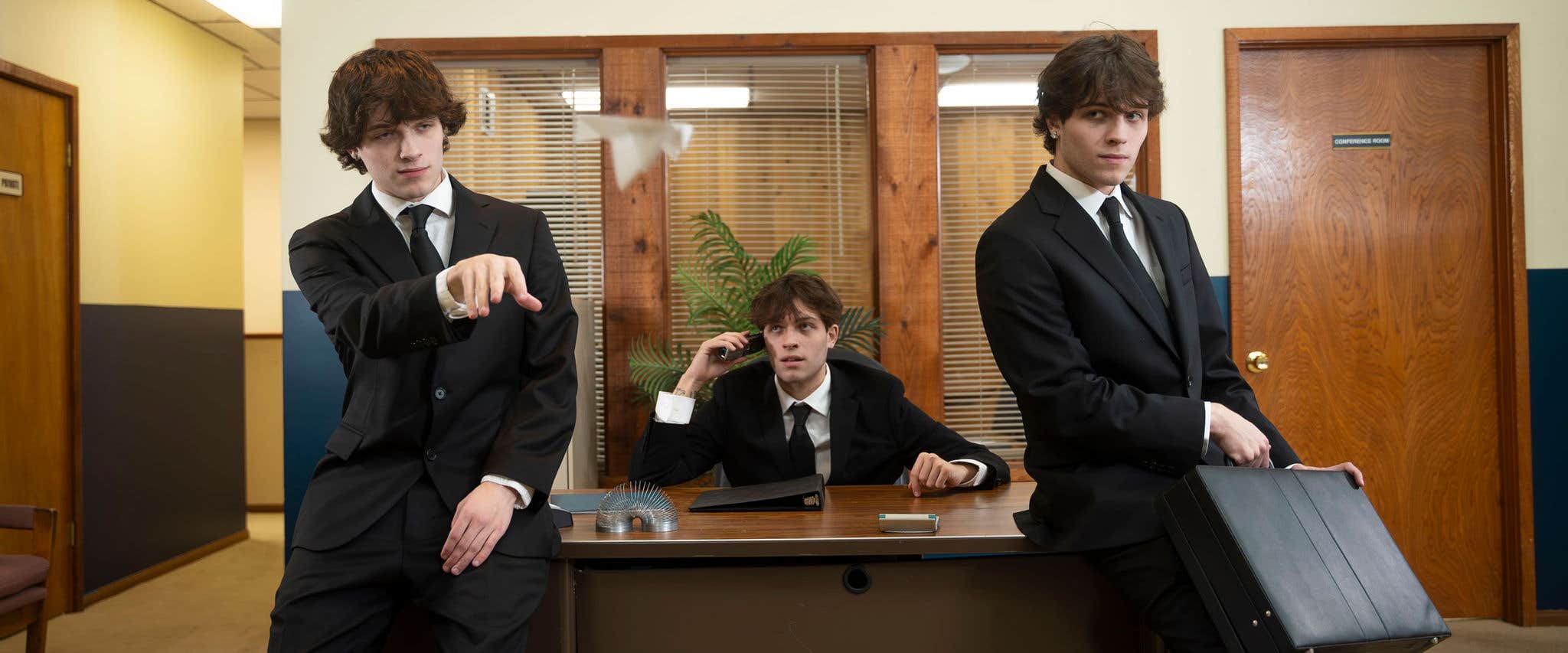 Sturniolo Triplets in suits in an office; one tossing a paper airplane, another on the phone, and the third holding a briefcase.