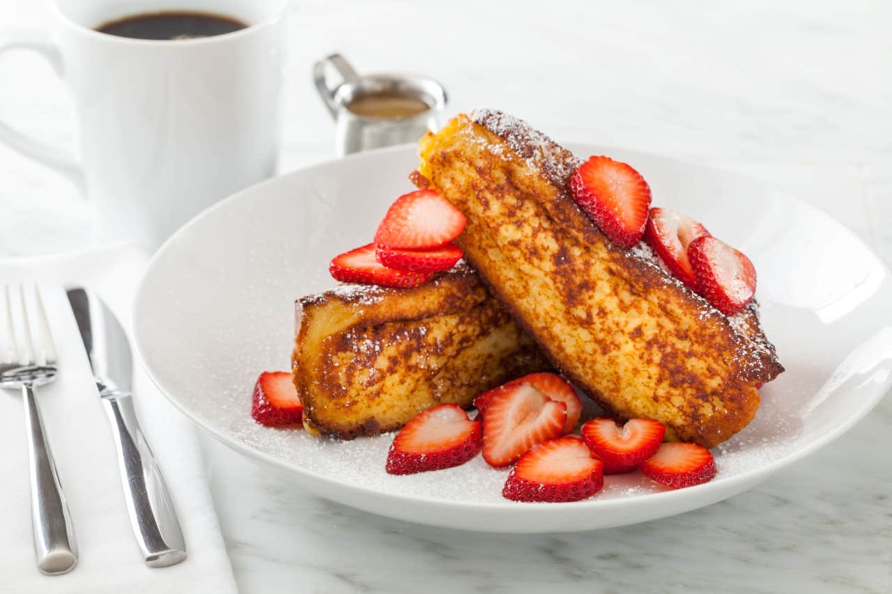 Thick slices of French toast topped with fresh strawberries and powdered sugar, served with coffee and cutlery nearby.