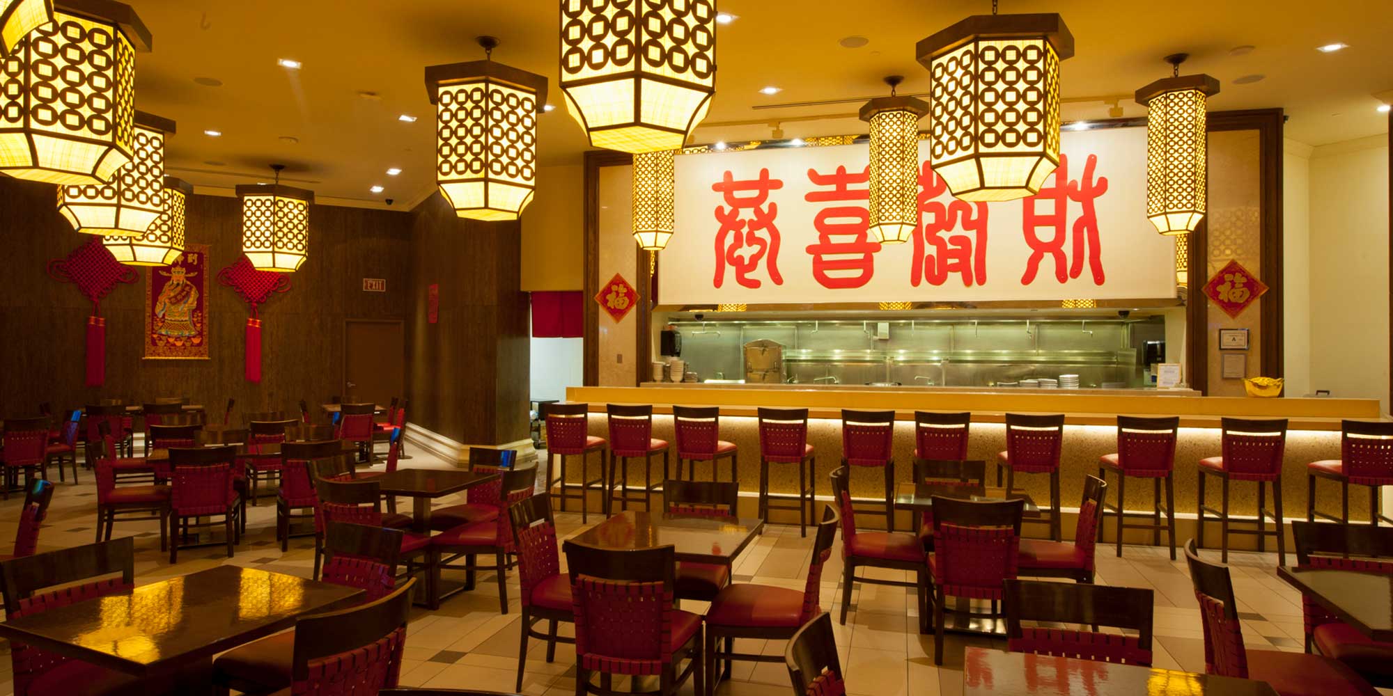 Empty restaurant with wooden tables and chairs, lantern-style lights hanging from the ceiling, and a large kitchen in the back.