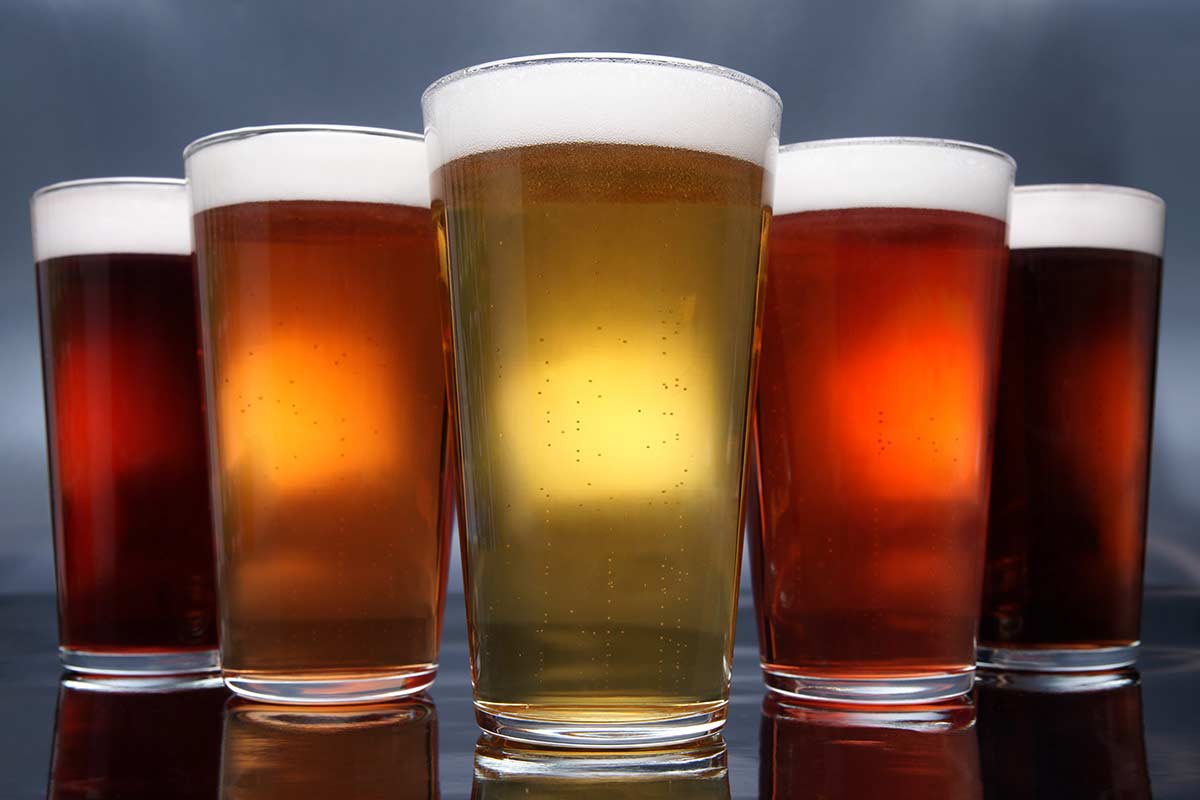 Five glasses of beer in varying colors, from dark brown to light amber, arranged in a row with foamy heads on top.