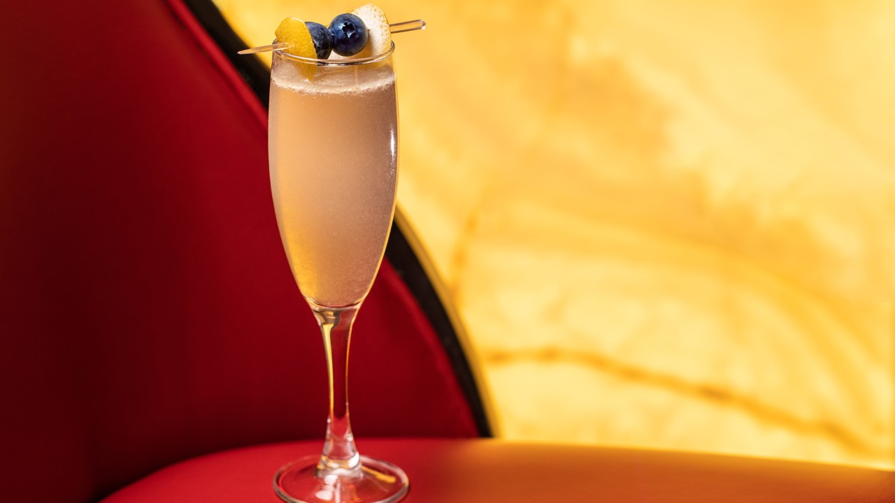 A sparkling cocktail garnished with a lemon twist and blueberries, served in a champagne flute against a warm, soft background.