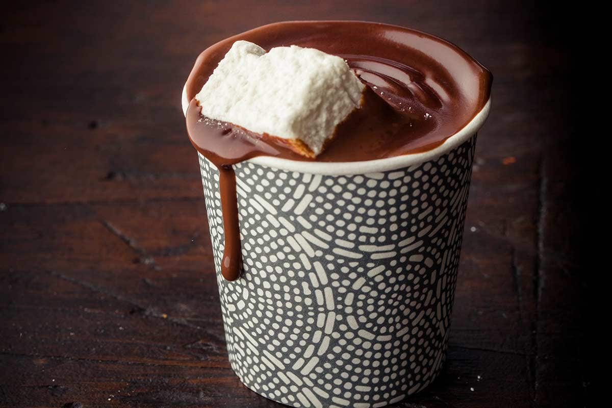 A cup of rich hot chocolate with a large marshmallow on top, overflowing slightly, on a dark wooden surface.