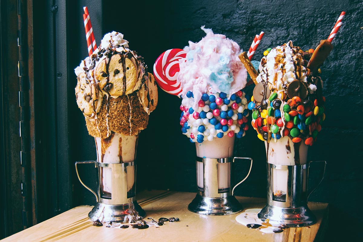 Three extravagant milkshakes topped with cookies, cotton candy, lollipops, and colorful candies on a wooden table.