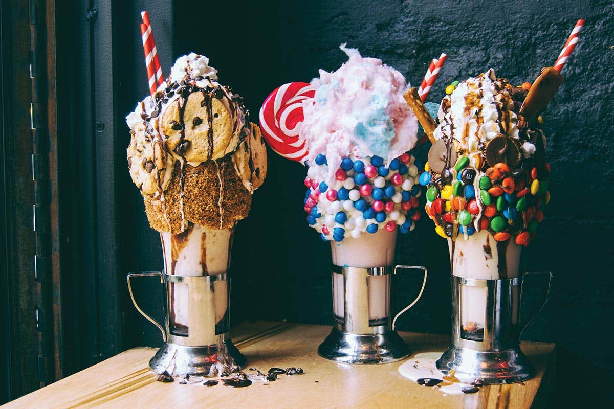 Three extravagant milkshakes topped with cookies, cotton candy, lollipops, and colorful candies on a wooden table.