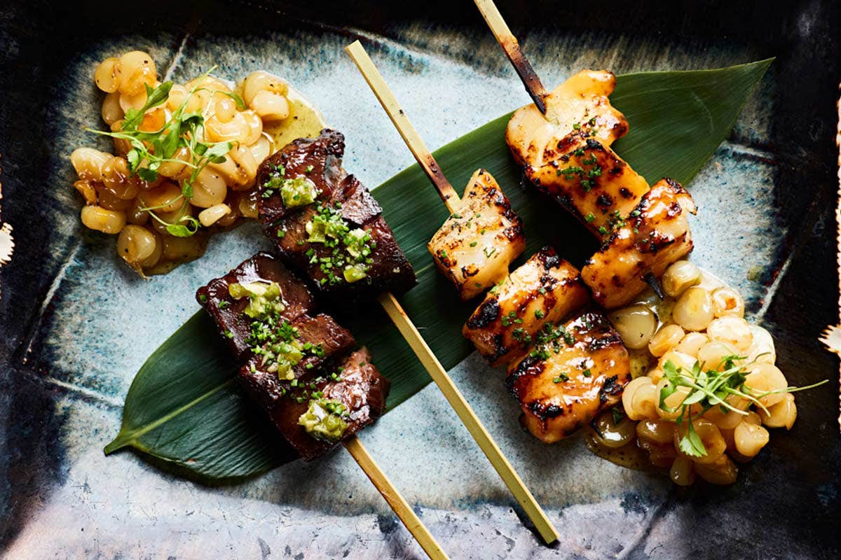 Three skewers of grilled meat and vegetables on a dark plate, garnished with fresh herbs and accompanied by cooked beans.