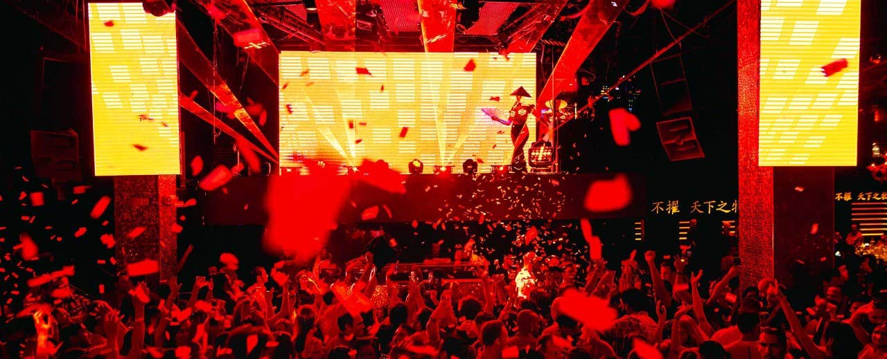 Crowd dancing at a lively nightclub with bright lights, falling confetti, and a large LED screen displaying visuals.