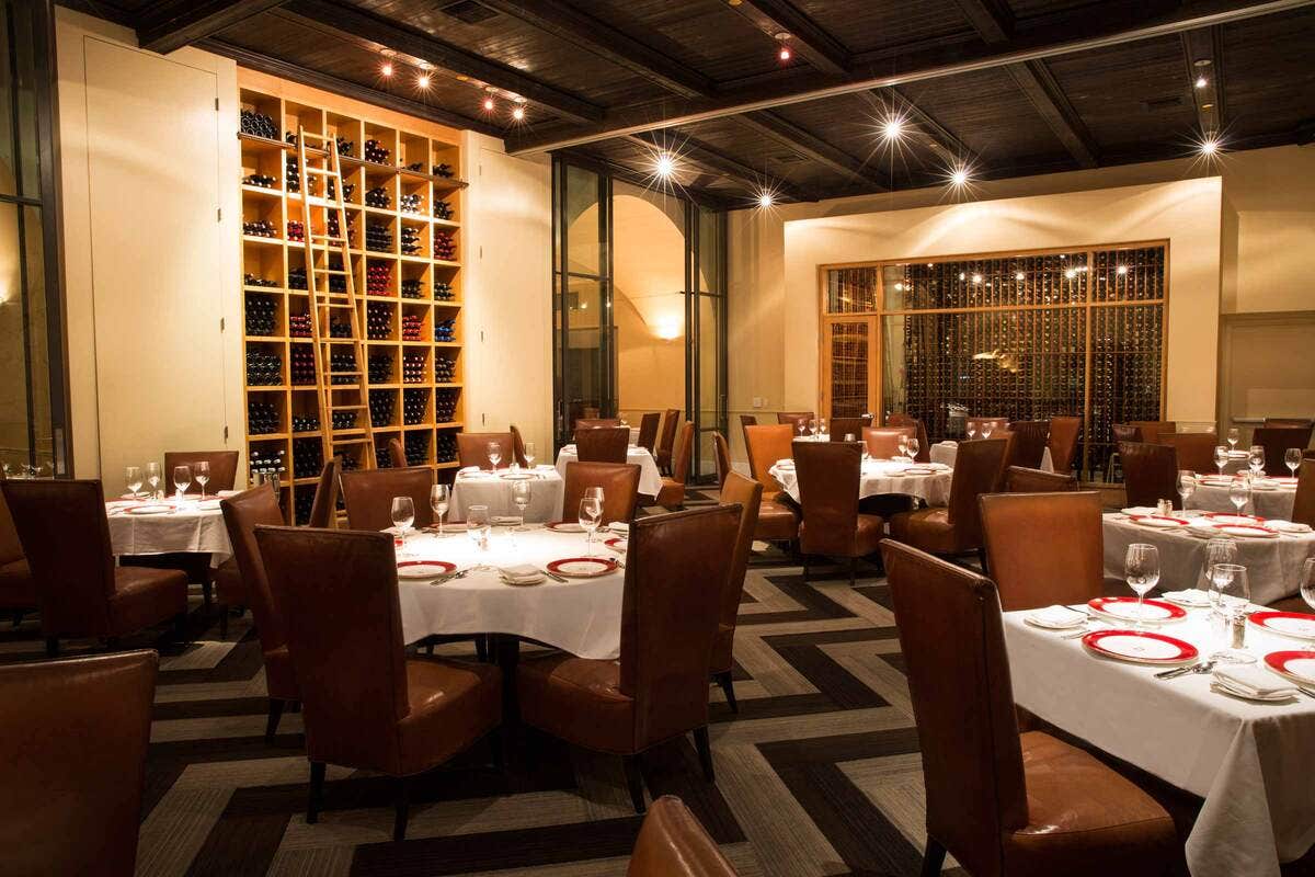 Elegant restaurant with round tables set with white tablecloths and brown leather chairs, and shelves stocked with wine bottles.