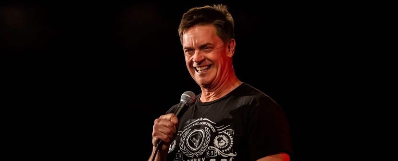 A man on stage smiling while holding a microphone, wearing a black T-shirt with a graphic design.