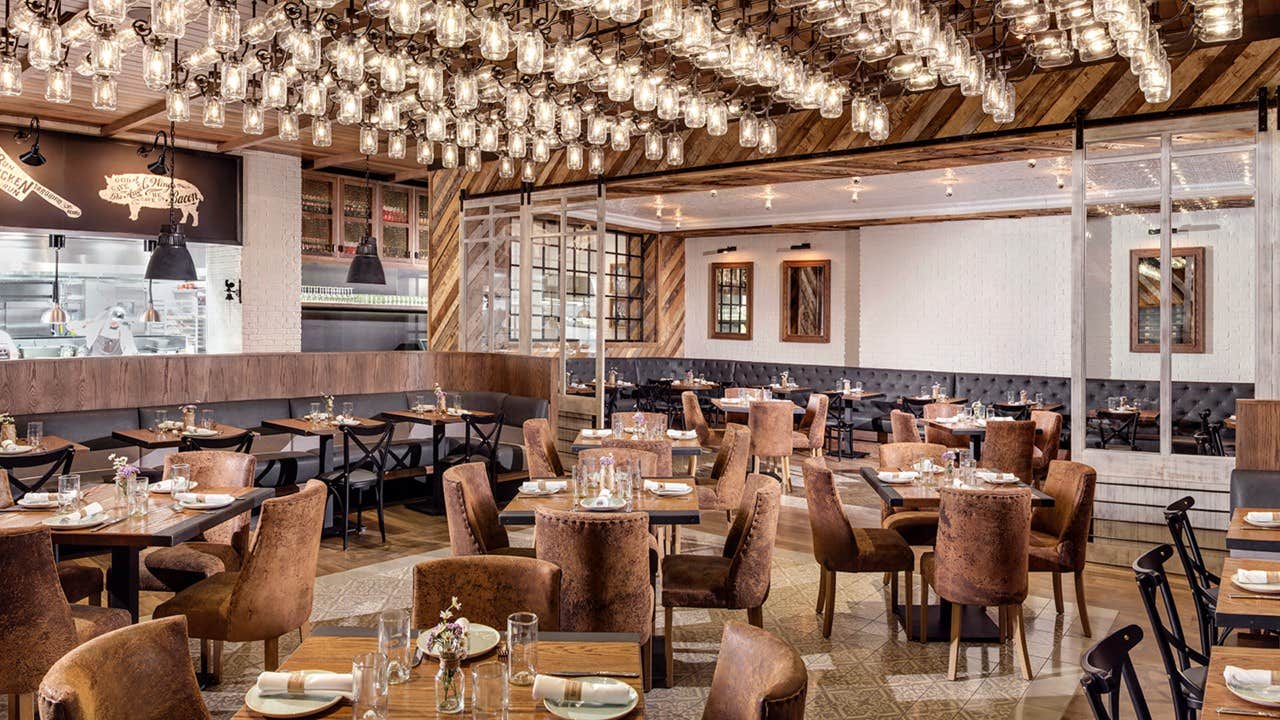 Elegant restaurant interior with wooden tables and plush chairs, featuring warm lighting and a glimpse of the kitchen on the left.