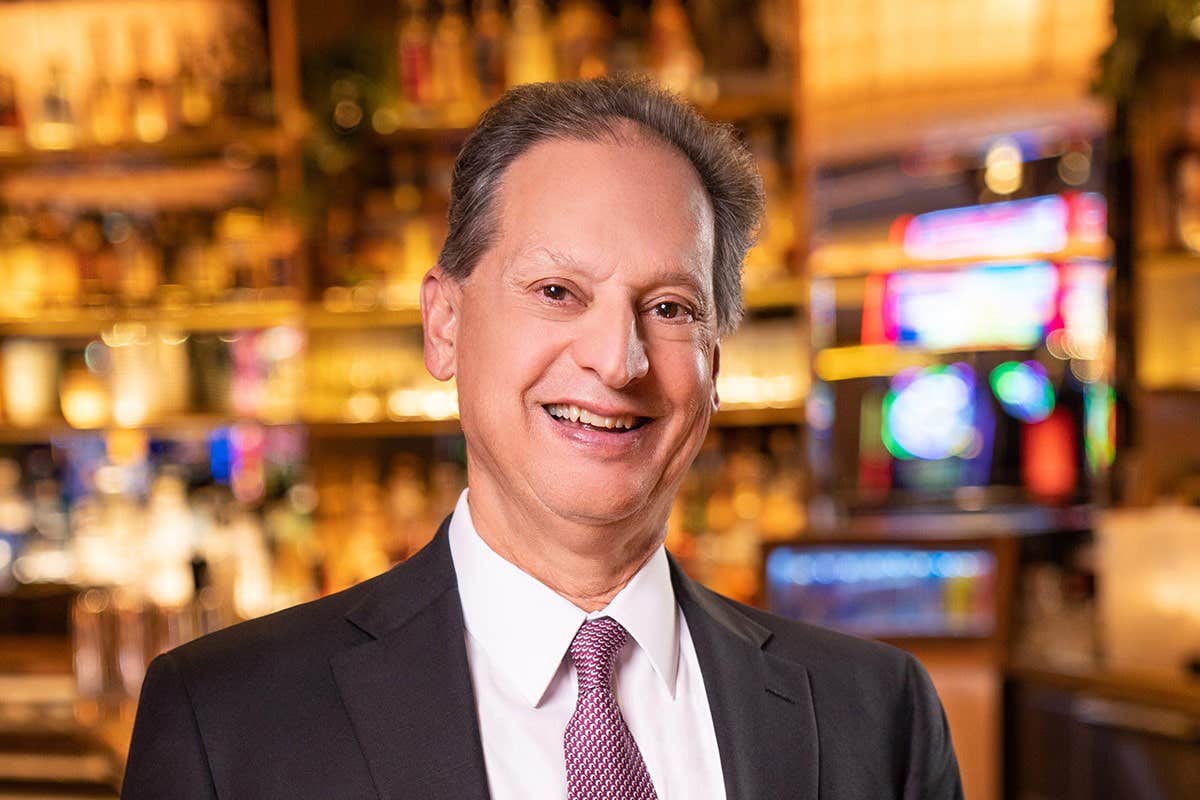 A man in a suit and tie smiles in a warmly lit room with a bar in the background.