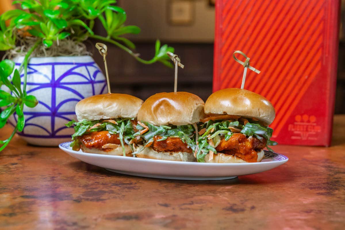 Three buffalo chicken sliders with lettuce on a white plate, placed on a table with a potted plant and red-striped box nearby.