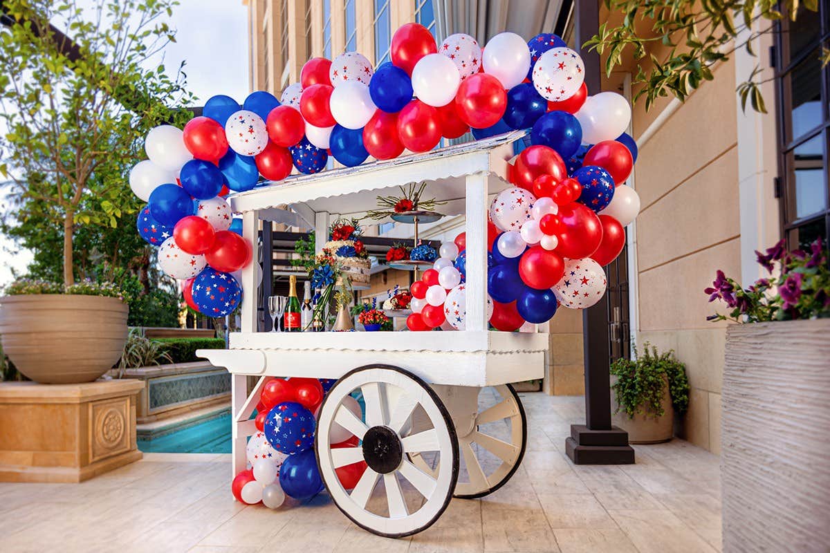 A decorated white cart with red, white, and blue balloons next to a swimming pool and building.