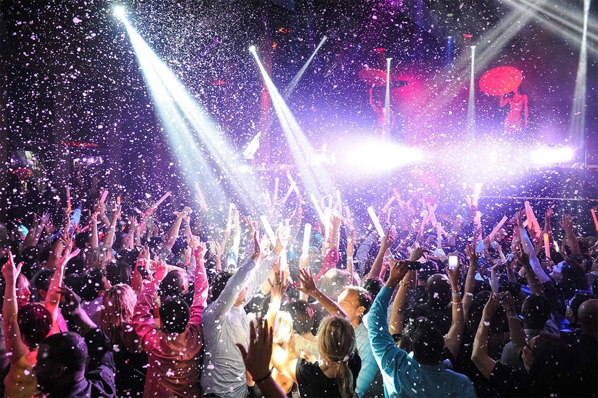 A crowded nightclub with people dancing under bright lights and snow-like confetti falling from above.