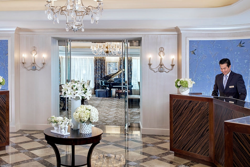 Elegant hotel lobby with two wooden reception desks, floral arrangements, and a chandelier. A grand piano is in the background.