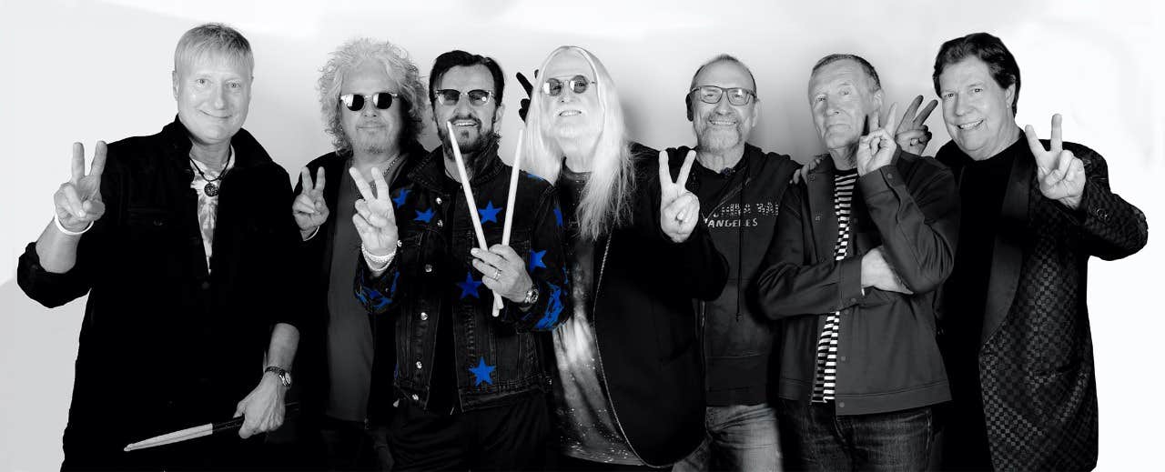 A group of seven men in casual clothing holding up peace signs and smiling, some with sunglasses, in a black and white photo.