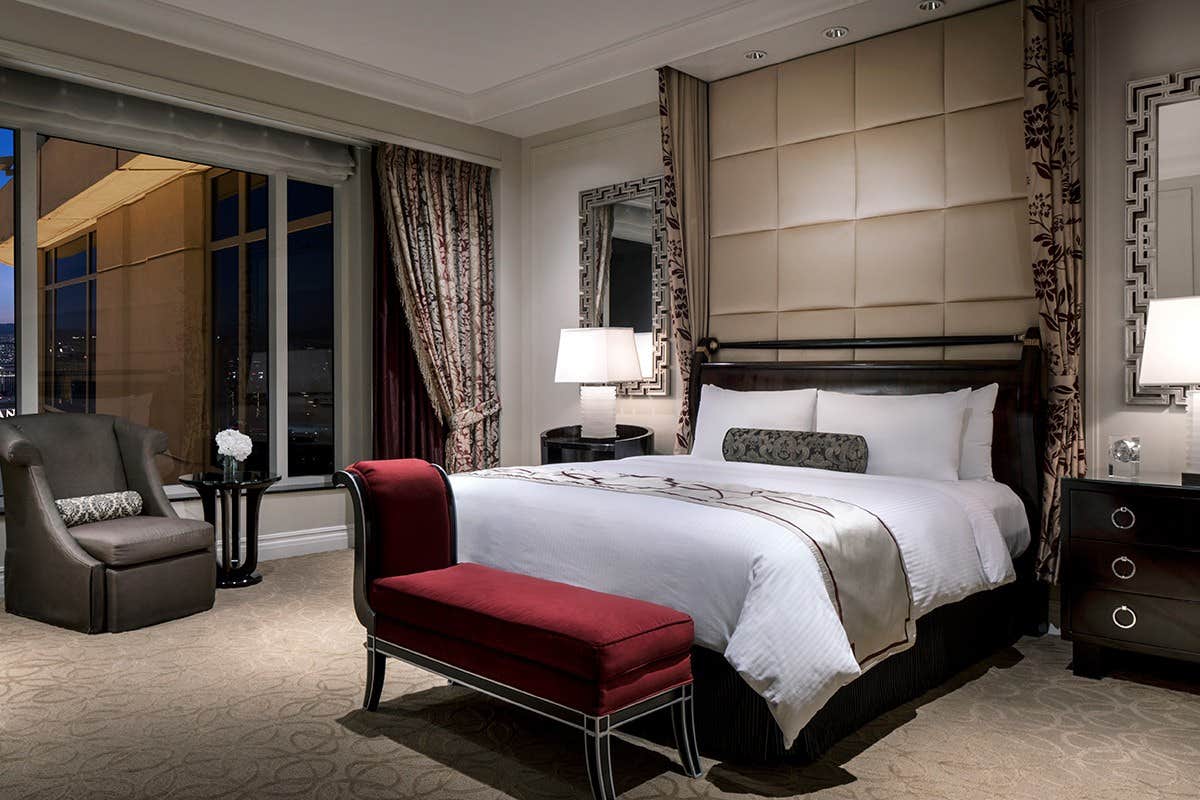 Luxury hotel room with a king-sized bed, red bench, armchair by the window, nightstand and elegant decor.