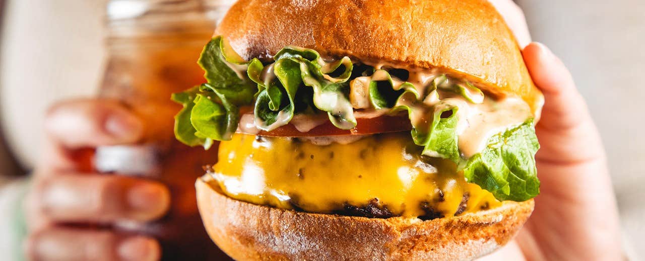 Close-up of a hand holding a cheeseburger with lettuce, tomato, and sauce, beside another hand holding a beverage in a jar.