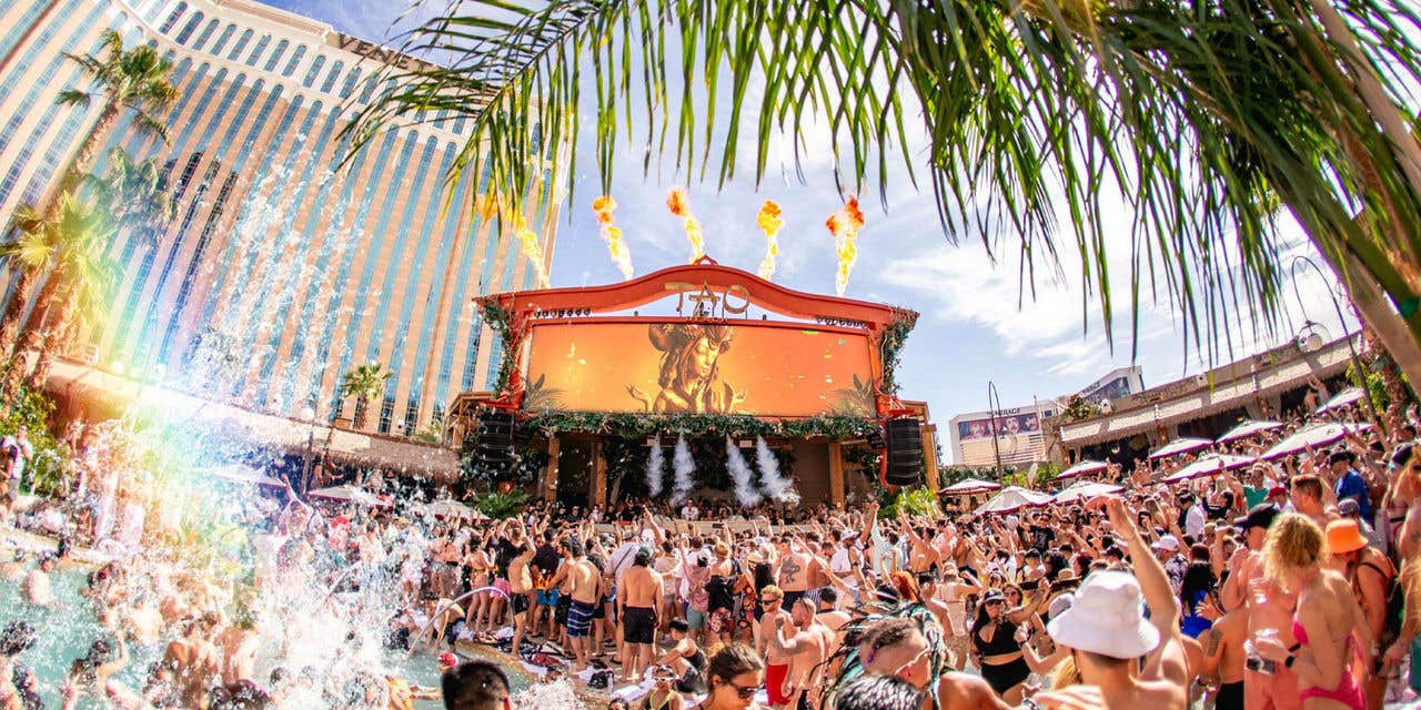 A large crowd at TAO Beach, with a rainbow refraction on the left from a water splash in the pool, and flames going up from behind the stage, the picture is framed with palm trees and The Venetian Resort in the background