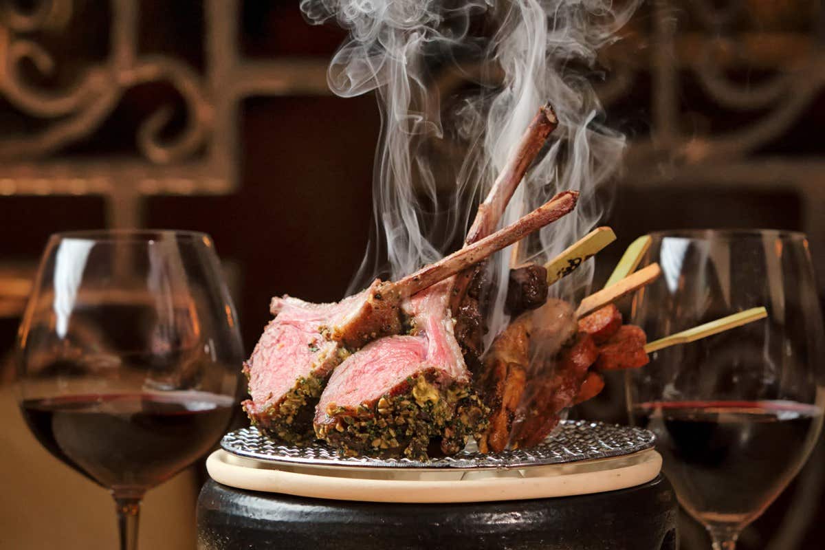 Two glasses of red wine beside a plate of steaming lamb chops and skewered meat.