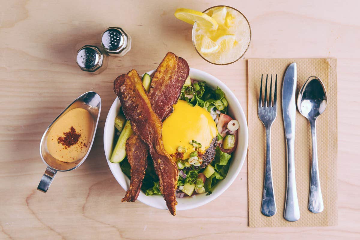 A bowl of salad topped with bacon and cheese, a glass of lemonade, utensils, and a sauce on a wooden table.