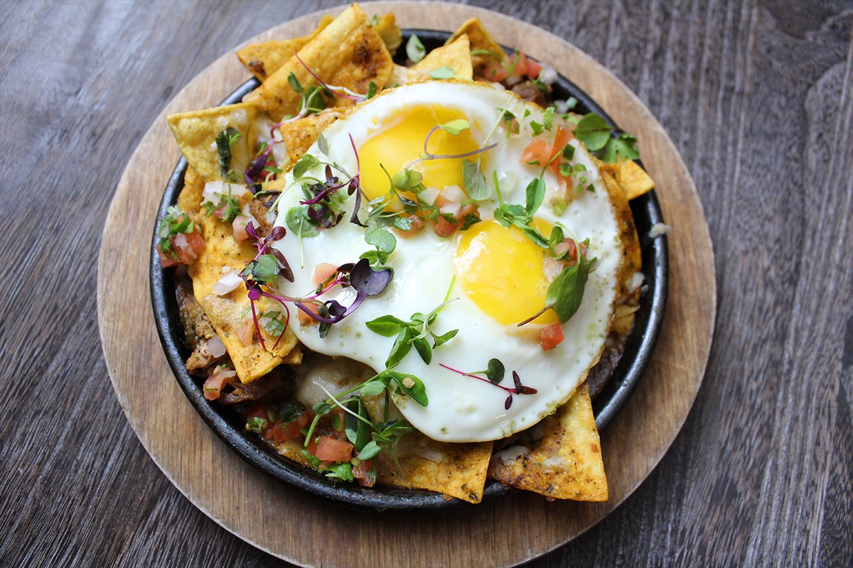 A plate of nachos with two sunny-side-up eggs, garnished with diced vegetables and greens, on a wooden surface.