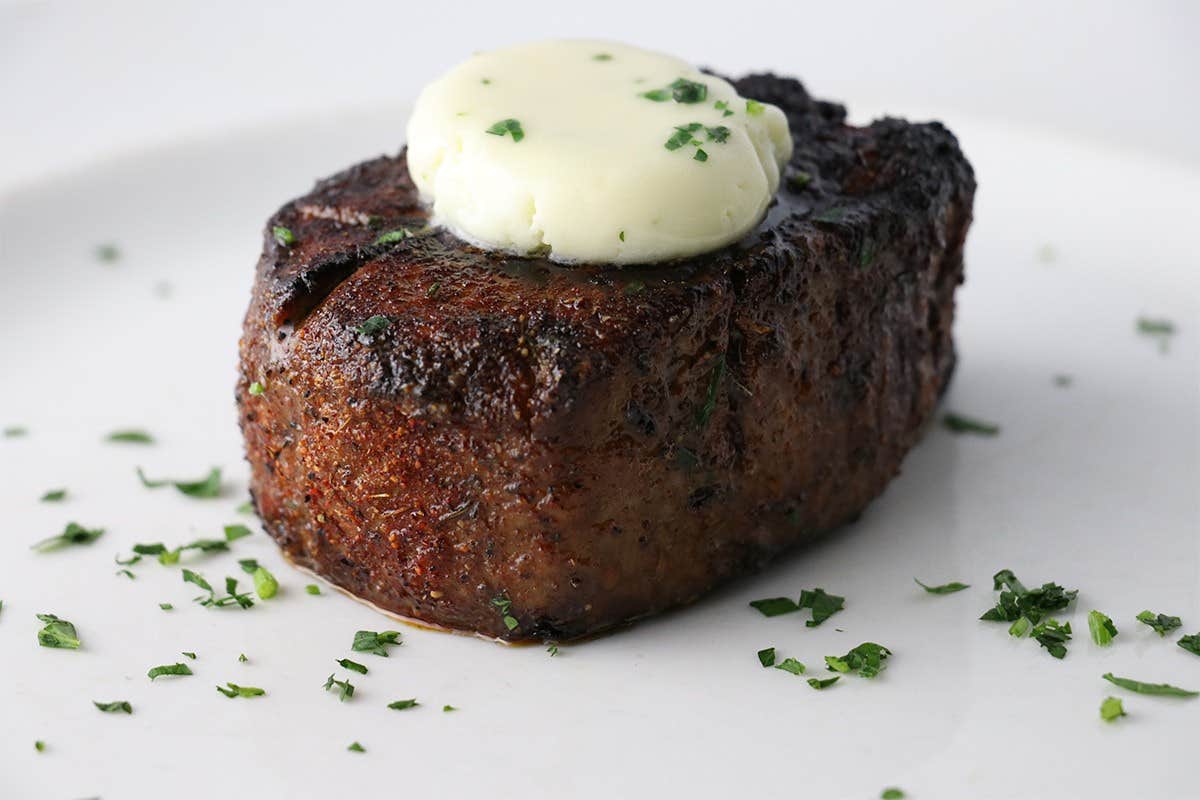 Grilled steak topped with a pat of melting butter and garnished with chopped herbs on a white plate.