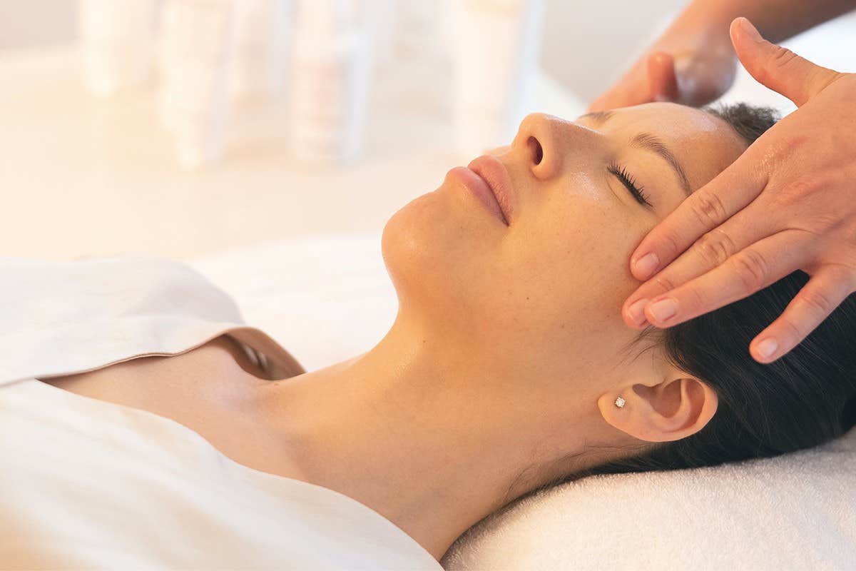 A person lying down with closed eyes receiving a relaxing facial massage from someone using both hands.