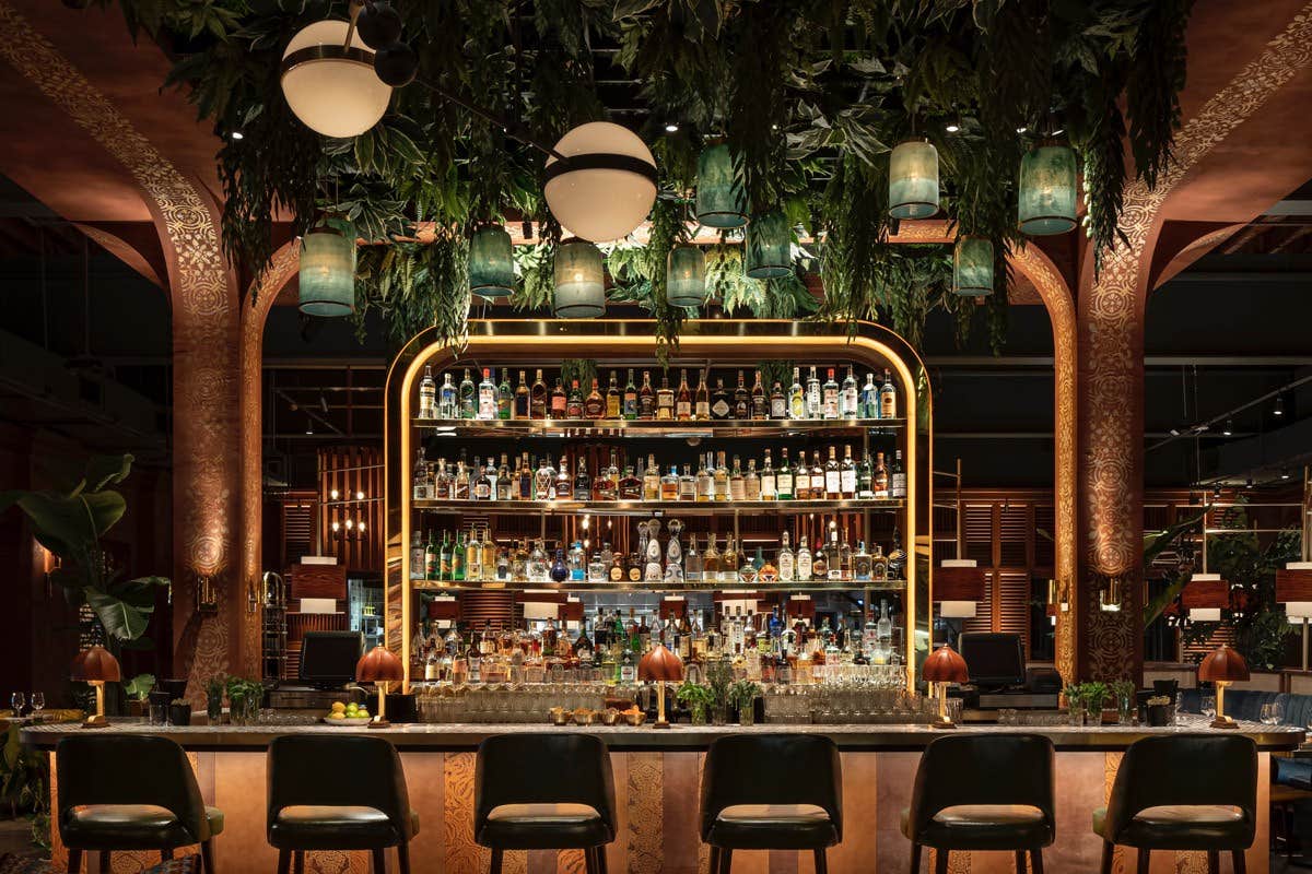 Chic bar with a lush green canopy, ambient lighting, a variety of drinks on shelves, and a line of elegant bar stools.