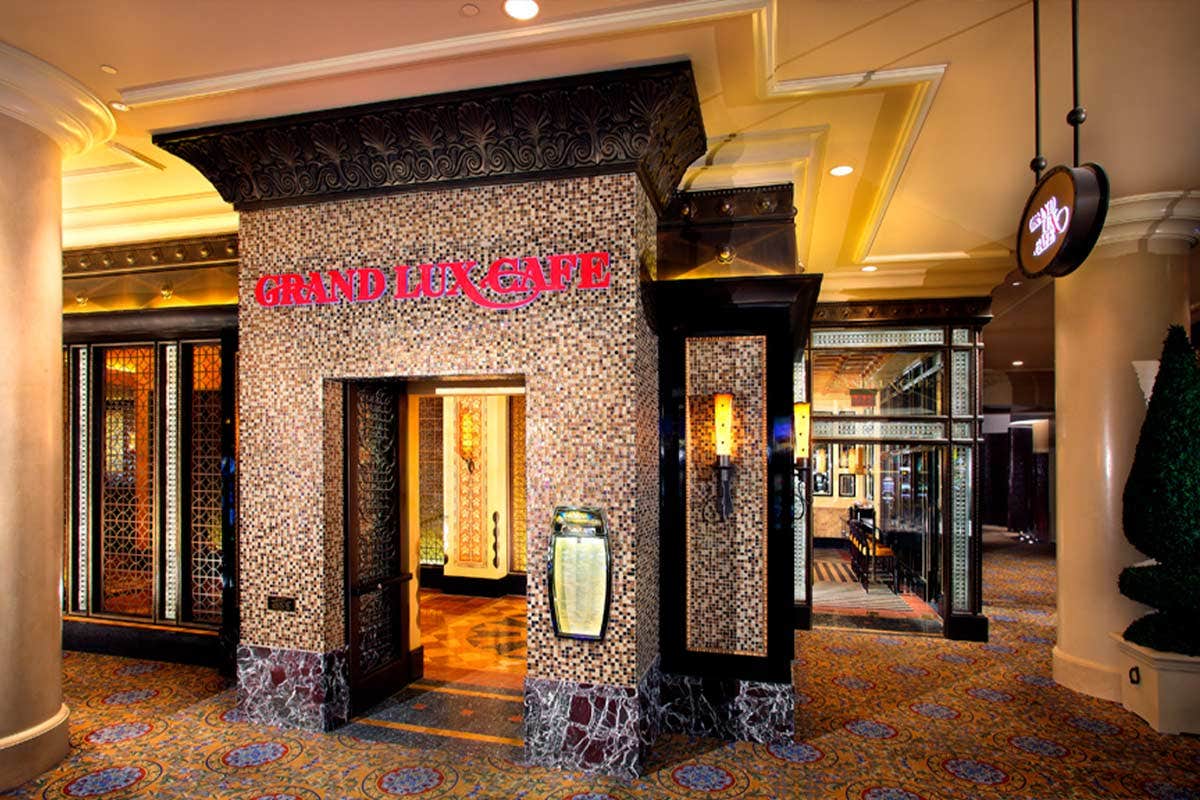 Entrance of Grand Lux Cafe with ornate, tiled design, framed by tall columns, and a small illuminated sign to the right.