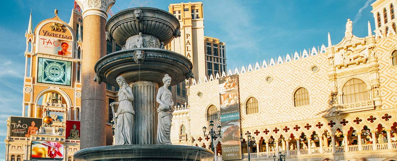 Decorative fountain with statues in front of ornate buildings, including casinos, in sunlight.