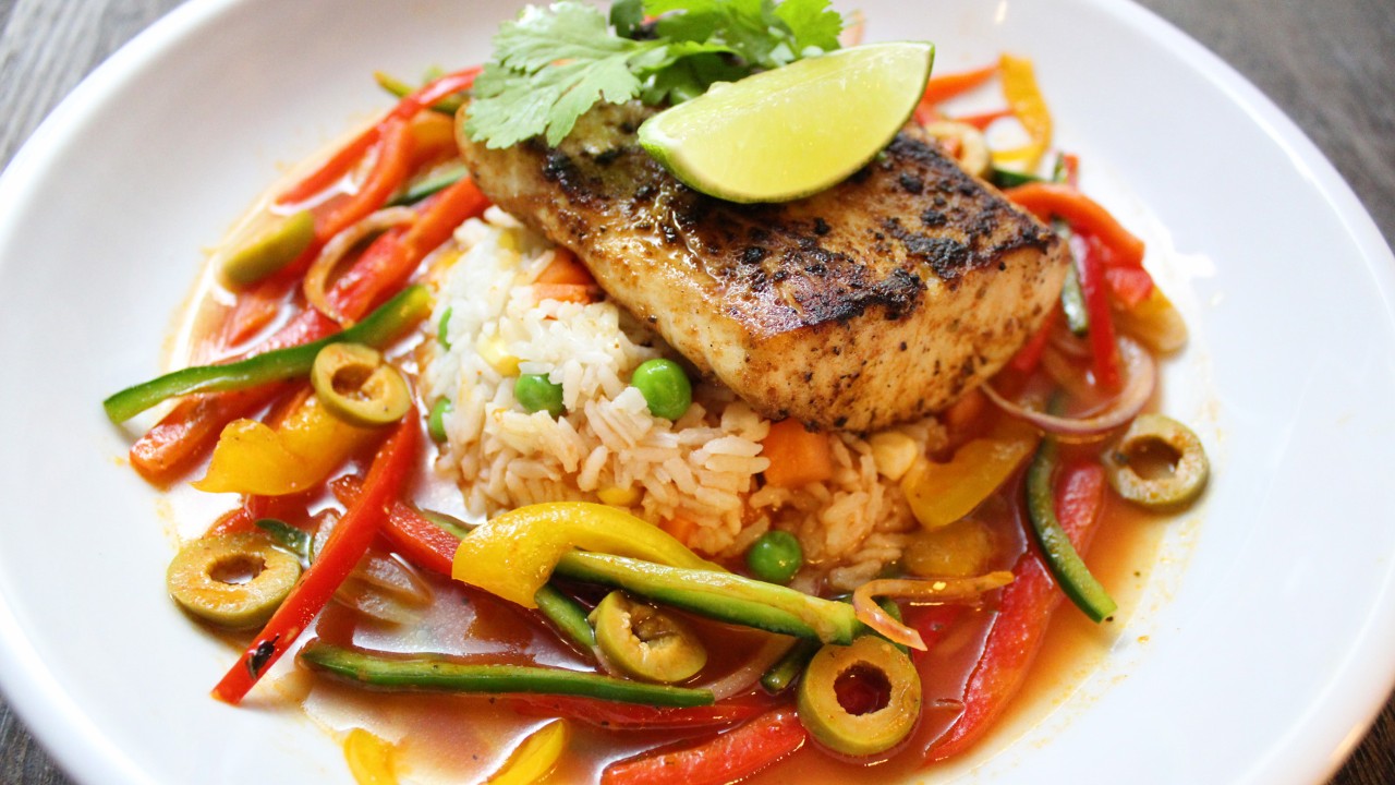 Grilled fish fillet on a bed of rice with colorful vegetables and a lime wedge on a white plate.