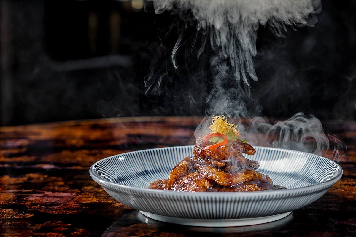 A steaming dish of glazed meat topped with thin sliced vegetables, resting on a white and blue-striped plate.