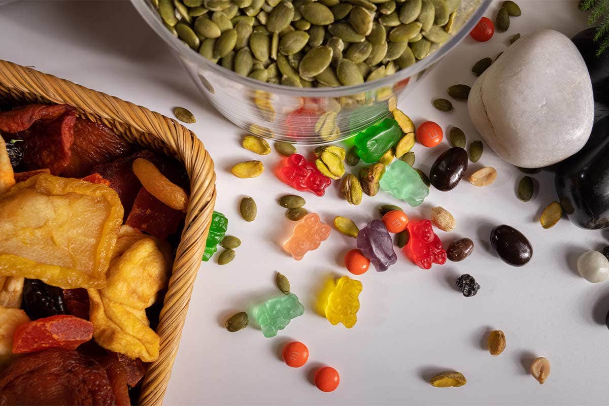 A mix of dried fruits, gummy bears, seeds, and candies scattered on a table, next to a container of pumpkin seeds.