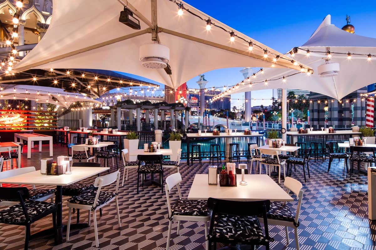 An outdoor restaurant patio at dusk with string lights, white tables, and chairs under large white canopy tents.