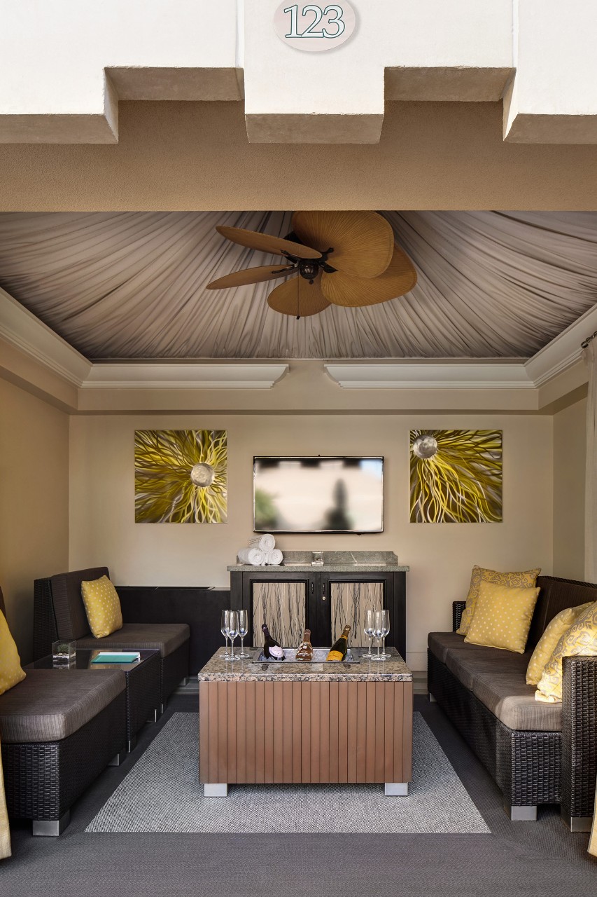 Outdoor cabana with wicker sofas, yellow cushions, coffee table, wall art, TV, and ceiling fan under a tented roof. Room number 123.