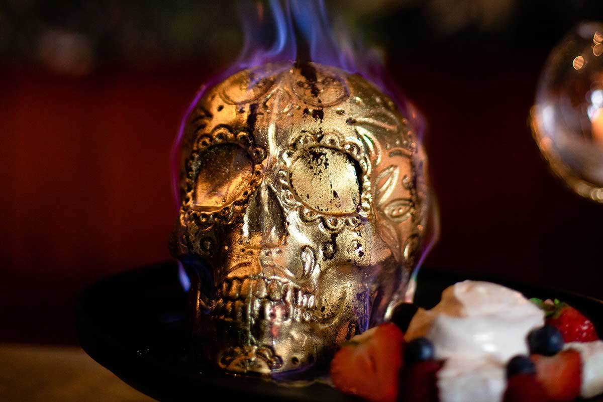 A decorated golden skull with purple flames on top, placed next to a dessert with whipped cream and berries.