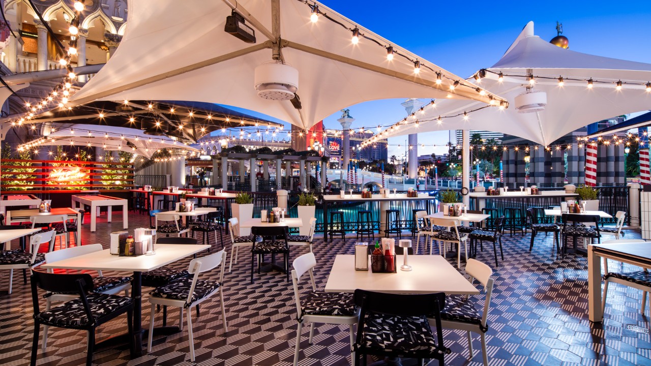 An outdoor restaurant patio with string lights, white tables, chairs, and a geometric tile floor at twilight.