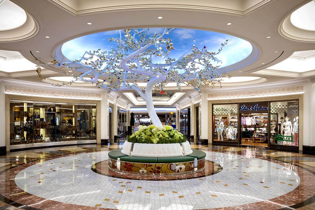 A luxurious indoor shopping area with a white tree sculpture and circular seating under a domed ceiling with a sky mural.