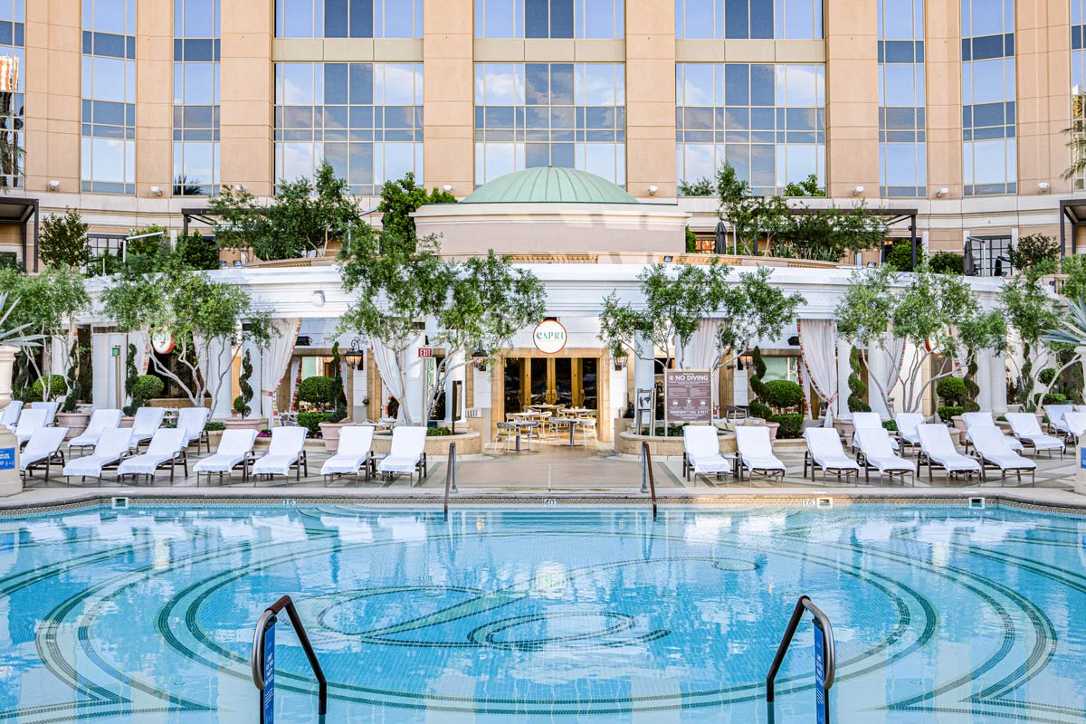 Opulent outdoor pool area with white lounge chairs, surrounded by trees and adjacent to a luxury building with large windows.