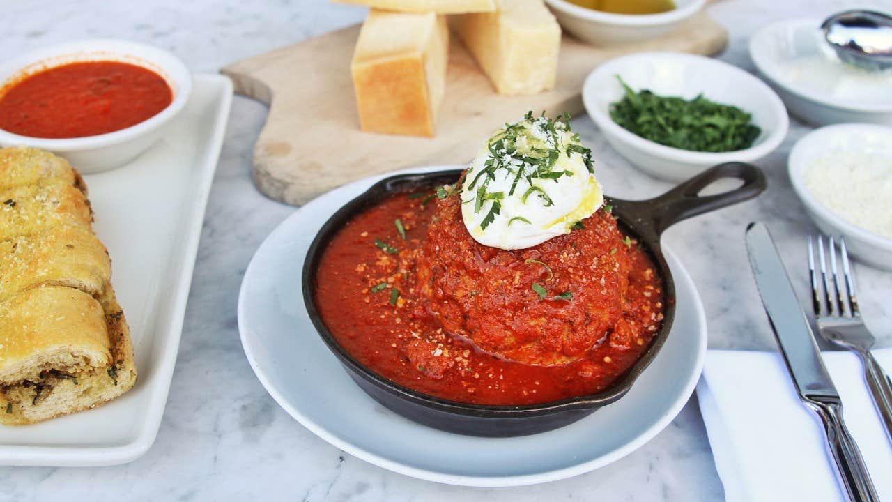 A skillet with a large meatball topped with ricotta on tomato sauce, surrounded by bread and small dishes on a table.